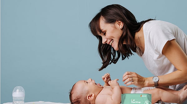 Person leaning over a baby lying on a changing mat in an indoor setting. A pack of baby wipes is visible in the foreground. The scene focuses on a caregiving moment between the person and baby, with a light blue background and soft natural lighting.